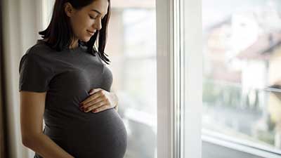 Young pregnant woman standing in front of window, holding belly